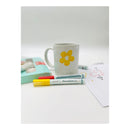 A white ceramic mug with a yellow flower design, styled on a white surface with two ZEBINE Percelain markers, a light blue box, and a piece of paper in the background.