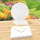 A stack of plain white paper envelopes sits on a wooden table with a blurred garden background. A single envelope is shown open behind it, revealing a circular backing card or insert.