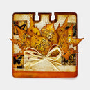 A square decorative card or art piece featuring layered autumn leaves in warm orange, yellow, and brown tones, with vintage-style text, butterfly motifs, and a cream twine bow at the center, shown on a light gray background.