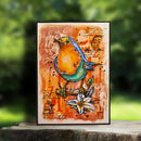 A framed mixed-media art print featuring a vibrant robin perched on a branch with a white lily flower, set against a warm orange and brown textured background with handwritten script details. The print is displayed on a rustic wooden surface with a blurred green garden background.