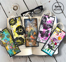 Five decorative paper bookmark tags with colorful mixed-media artwork are arranged on an open book on a whitewashed wooden surface. A pair of black eyeglasses rests above the book, and a circular logo in the top right reads 'llana creative designs by anna lisa esposito'.