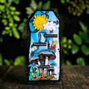 A decorative rectangular card or plaque with a black scalloped border, featuring a whimsical illustrated design of blue mushrooms, a smiling yellow sun, a white cloud, a bee, and the words 'Adventure', 'Experience', and 'Journey'. The item is photographed standing upright on a dark wooden surface with a blurred green garden background.