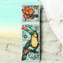 A vertical rectangular stamp or card design featuring a colorful toucan and orange flower on a distressed teal background, with a white scalloped border, shown against a beach-like backdrop with turquoise water and pale sand.