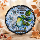 A circular mixed-media art print featuring a blue and green bird perched among white flowers and green leaves on a distressed blue background with the text 'Life's good, you should get one'. The print is displayed on a rustic wooden surface surrounded by autumn leaves, small red berries, and a soft beige fabric.