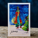 A vertical greeting card featuring a festive watercolor illustration of a woman in a red and green winter outfit walking beside a decorated Christmas tree under a snowy blue sky. The card has a white border and the word 'Xmas' written in black script at the bottom, displayed on a wooden surface with a dark blue background.