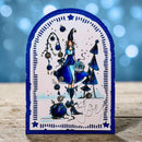 A handcrafted arch-shaped Christmas card featuring an illustrated woman in a blue dress and hat beside a decorated tree, with the word 'Joy' in white script at the bottom, photographed on a wooden surface against a blue bokeh background.