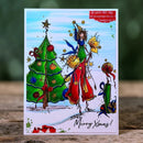 A handmade Christmas greeting card featuring a whimsical illustration of a woman in red and green clothing dancing beside a decorated Christmas tree, with the text 'Merry Xmas!' at the bottom. The card is displayed upright on a wooden surface with a blurred green background.