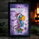 A rectangular black-edged card or plaque featuring a colorful Christmas parrot illustration on a distressed purple background with the text 'Merry Christmas' and '507'. The item is photographed on a rustic surface with a blurred warm fireplace and evergreen branches in the background.