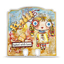 A decorative scrapbook card or craft plaque featuring a whimsical illustrated girl with braided pigtails, a gingerbread house, holiday decorations, and the phrase 'baked with love' on a textured beige and blue background.