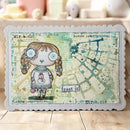 A rectangular mixed-media art card with scalloped edges, featuring a whimsical illustrated girl with braided pigtails, blue eyes, and a snowman sweater, alongside a distressed map design and text including 'frost it' and 'Miracle.' The card is photographed standing on a light wood surface with a softly blurred indoor background.
