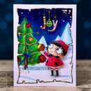 A handmade Christmas greeting card featuring a whimsical illustration of a girl in a red coat decorating a green Christmas tree under a dark blue night sky, displayed on a wooden surface with a dark blue bokeh background.
