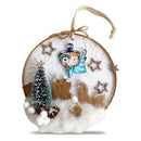 A circular embroidered hanging ornament featuring a handmade winter village scene with a small angel figure, decorated wooden houses, a miniature evergreen tree, stars, and faux snow on a white embroidered background.