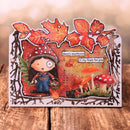 A handmade layered card featuring a whimsical cartoon girl in overalls under a red-and-white mushroom hat, surrounded by autumn leaves, mushrooms, and the text 'there's mushroom in my heart for you', displayed on a wooden surface.