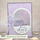 A handmade greeting card with a lavender background, an oval white center featuring a winter tree scene and the words 'GOOD cheer TO YOU', a sheer white ribbon bow, and a decorative border. The card is photographed on a wood surface with a neutral wall behind it.