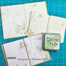 A styled flat-lay of handmade scrapbook journals with stamped and painted details, alongside an open ink pad labeled 'Distress Oxide' by Arden Creations Studio. The journals feature beige and white textured paper with green and brown sketch-like designs.