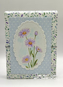 A handmade paper journal or scrapbook box featuring a light blue polka dot background with a central white scalloped oval containing painted purple daisies with yellow centers. The sides have a floral border pattern in green and purple.