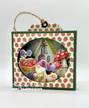 A square decorative hanging plaque with a green border and strawberry-patterned backing, featuring a circular cutout of a whimsical cottage scene with a gnome, mushrooms, strawberries, and flowers. A twine loop hangs from the top, and a watermark reading 'Simone Carr ART IMPRESSIONS' appears near the bottom.