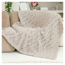 A chunky knit throw blanket in a light beige color with an openwork leaf pattern, draped over a beige armchair in a cozy living room setting.