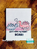 A handmade greeting card featuring two cartoon dinosaurs, one blue-gray and one pink, with a small heart above them. The card reads 'you make my heart ROAR!' in black lettering.