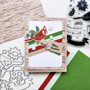 A handmade Christmas greeting card featuring a red cardinal, green and red stripes, snowflakes, pine branches, and a kraft paper tag that reads 'Merry Christmas.' The card is styled on a tabletop with crafting supplies, including clear stamps, metal dies, patterned paper, and colored cardstock.