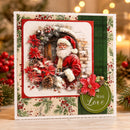 A square handmade Christmas greeting card displayed on a wooden surface with a festive blurred background. The card features a central illustration of Santa Claus surrounded by a wreath, red poinsettias, pinecones, and a brick wall, with decorative borders and a green circular badge that reads 'With Love'.