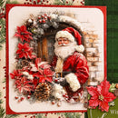 A square Christmas card featuring a festive 3D-style illustration of Santa Claus in a snowy arched doorway, surrounded by a wreath of red poinsettias, pinecones, berries, and snow. The card has a red border and is photographed on a decorative holiday-themed surface with green plaid and floral elements.