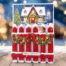 A Christmas greeting card or decorative sign featuring a snowy house, decorated fence, evergreen tree, ornaments, and a bird, with the text 'Merry Christmas' at the bottom, shown against a blurred festive background.