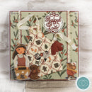 A square handmade scrapbook card featuring a rustic baking theme, with a central cream apron patterned with red and brown motifs, a white lace circle reading 'Baked Love,' illustrated characters, and a green floral border on a light fabric background.
