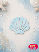 A light blue seashell-shaped craft die or stencil with fan-shaped cutouts, displayed on a textured white and blue background with decorative seashells, coral, and clear water droplets. A 'CRAFT & YOU DESIGNS' logo is visible in the bottom right corner.