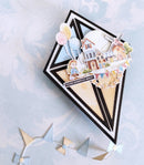A handmade layered card or box with a black and white geometric frame, decorated with pastel balloons, a village house, a windmill, and a small character figure. The card is placed on a pale blue marbled surface with a bunting garland nearby.