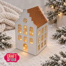 A small decorative house-shaped craft ornament with a warm glowing light inside, styled on a soft cream surface with snowy greenery and string lights around it. A pink 'CRAFT & YOU' logo appears in the lower left corner.