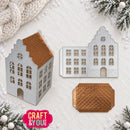 A holiday-themed product photo showing a metal die-cut craft template for a house, displayed both as a finished layered cutout with a copper-gold roof and as the flat metal stencil, plus a separate matching copper-gold textured piece. The background is a textured cream surface decorated with a chunky knit blanket, snowy pine branches, and white ornaments.
