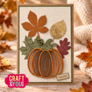 A handmade autumn-themed card featuring a layered paper pumpkin with orange and brown stripes, green and brown leaves, and a small 'Autumn' label, displayed on a rustic wooden surface with decorative fall leaves and a knitted blanket in the background. A 'CRAFT & YOU DESIGN' logo is visible in the bottom-left corner.