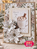 A handmade holiday greeting card featuring a central photo of a mug filled with hot cocoa and marshmallows, surrounded by layered cream poinsettia and snowflake embellishments, on a patterned background with a 'Warm & Cozy' design. A pink 'CRAFT & YOU' logo is visible in the bottom right corner.