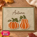 An autumn-themed handmade greeting card featuring two layered orange pumpkins with green glittery stems and leaves, the word 'Autumn' in brown script, and a kraft paper border. The card is displayed on a wooden surface with autumn leaves and burlap fabric in the background.