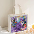 A clear plastic tote bag with white handles, featuring a colorful printed illustration of a purple cat surrounded by flowers and butterflies on the front, photographed in a softly lit indoor setting.