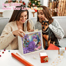 A woman holds a decorative gift tote bag featuring a purple cat illustration, with another woman and a small dog sitting beside her on a couch. The scene is decorated for Christmas, with wrapping paper and a small wrapped box on the table.