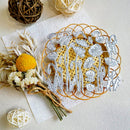 A floral metal cutting or stamping die set with white metal outlines of flowers and leaves over a gold patterned circular backing, styled on a textured white surface with dried flowers, twine balls, and natural twr.