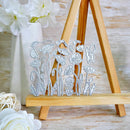 A silver metal die-cut floral stamp or craft template with poppy and dandelion shapes, displayed on a small wooden easel stand with decorative white flowers in the background.