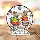 A round decorative holiday sign featuring two kissing mice in festive clothing, displayed on a wooden surface with a blurred warm bokeh background. The sign reads 'MERRY KISS-MOUSE' on its stand.