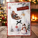 A handmade Christmas greeting card displayed on a wooden surface with a blurred holiday background. The card features a red border, a winter scene with a snowman, a child in a sled, and a snowflake design.