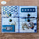 A handmade scrapbook album or journal spread photographed on a wooden surface. The album has a vintage industrial theme with blue and white tones, featuring hexagonal patterns, stars, a ticket stub, a speedometer graphic, and brass-tone buttons.