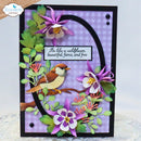 A handmade layered card featuring a bird, purple wildflowers, green leaves, and a black arch on a lavender gingham background, with a quote in the center. The card is framed in black and photographed against a light background.