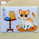 A handmade layered paper craft card featuring a cute orange and white cat sitting in an orange bowl, next to a small table with a blue can and a bag of cat food. The card has the text 'Life is BETTER with a CAT' and is displayed on a beige textured fabric background.