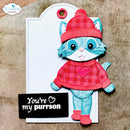A handmade paper craft tag featuring a cute layered paper cat wearing a red and pink plaid sweater and matching hat, with the text 'You're my purrson' on a black tag below, displayed on a textured brown background.