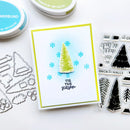 A handmade Christmas greeting card with a green pine tree and blue snowflakes, styled with crafting tools and ink pads on a white surface.