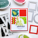 A handmade greeting card featuring a festive Christmas tree design and the words 'DECK THE HALLS,' styled on a white surface with craft supplies including ink pads, clear stamps, and metal frames.