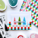 A flat-lay of a handmade Christmas card featuring three colorful Christmas trees, surrounded by crafting supplies including ink pads, patterned paper, and clear stamps on a white background.
