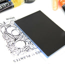 A black rectangular card or mat with a matte finish, shown resting on top of a white printed sheet with botanical line art. Partial colorful pens or markers are visible along the top edge of the frame.