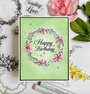 A handmade square greeting card with a light green background, featuring a floral wreath design with pink flowers and the words 'Happy Birthday' in black script. The card is styled on a white surface with craft stamps, colored markers, a pink rose, and green leaves.